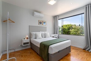 A modern bedroom with a bed, bedside table, and a view of greenery through large windows. - 2 km From Praia da Alvorada Near Barra Shopping Am203 (Rio de Janeiro)