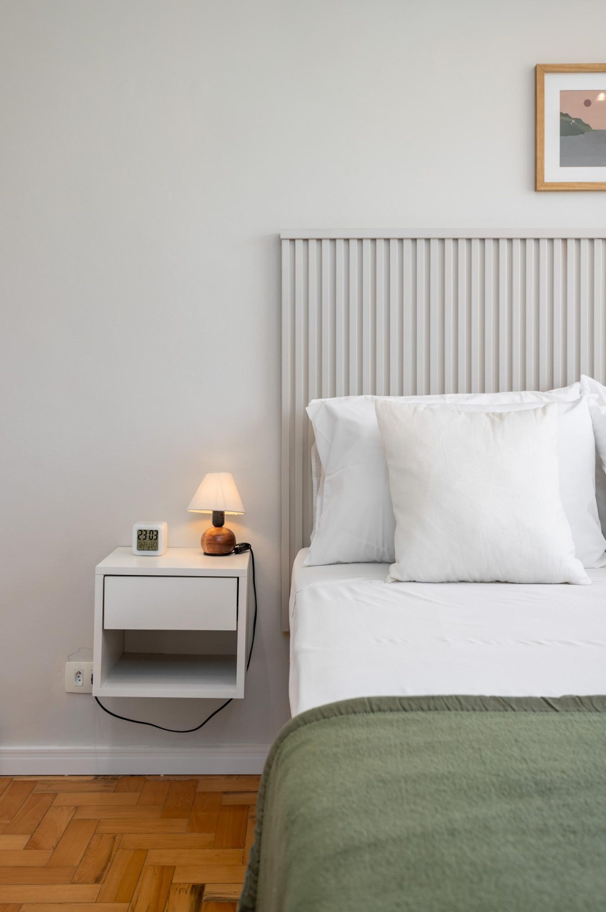 Un dormitorio ordenado con cama, mesita de noche con lámpara y un cuadro colgado en la pared.