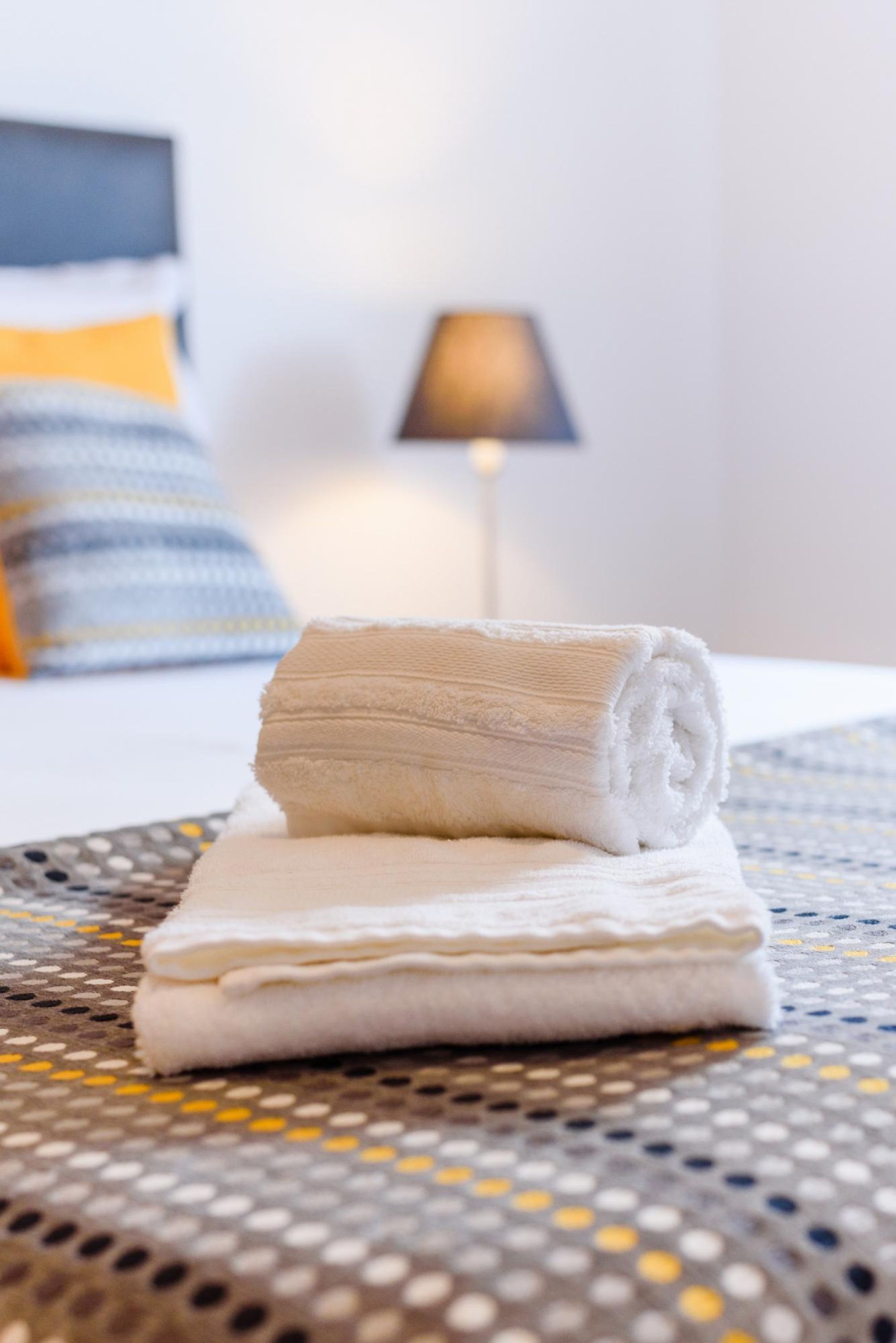A neatly folded towel on a bed with a patterned bedspread and a lamp in the background.