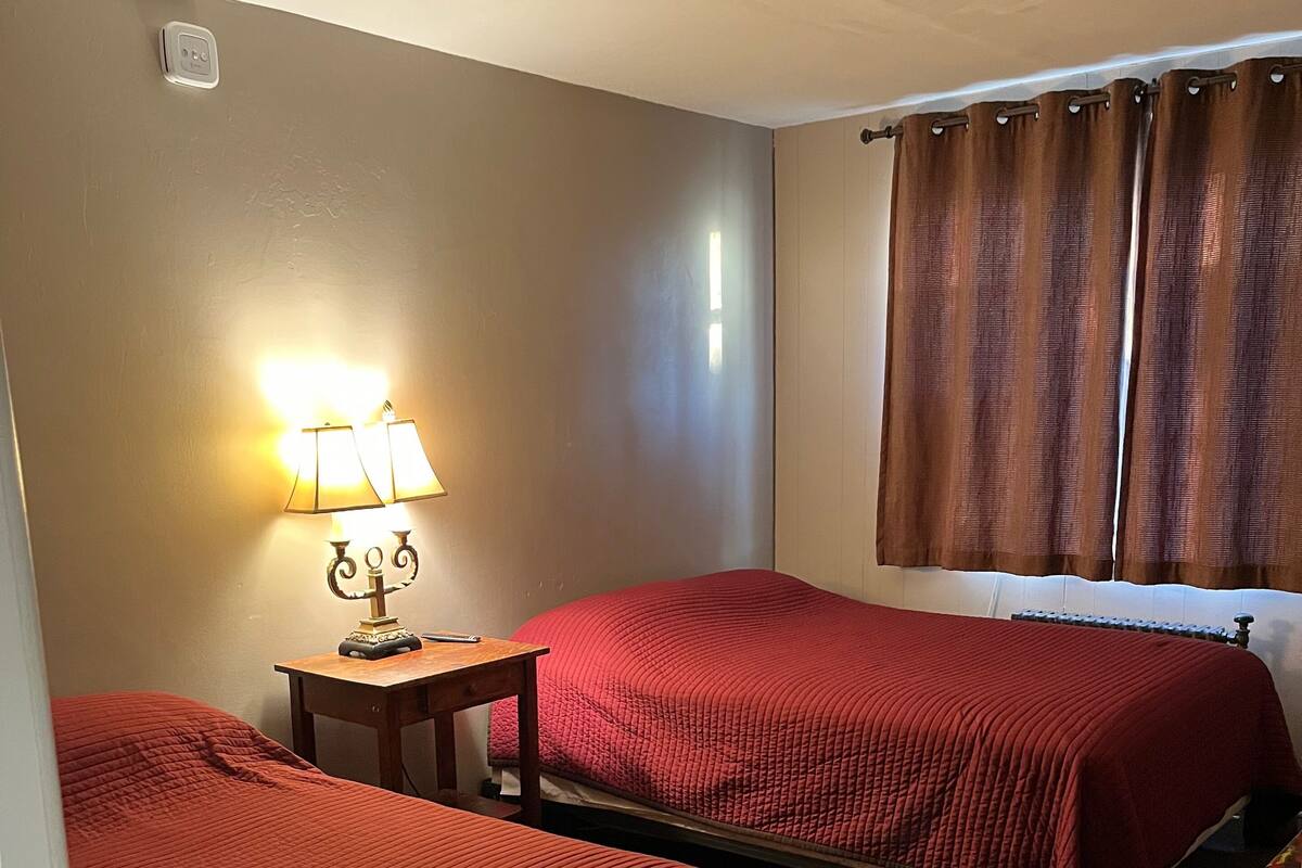 A room with two beds, a small table with a lamp, and a window with curtains.