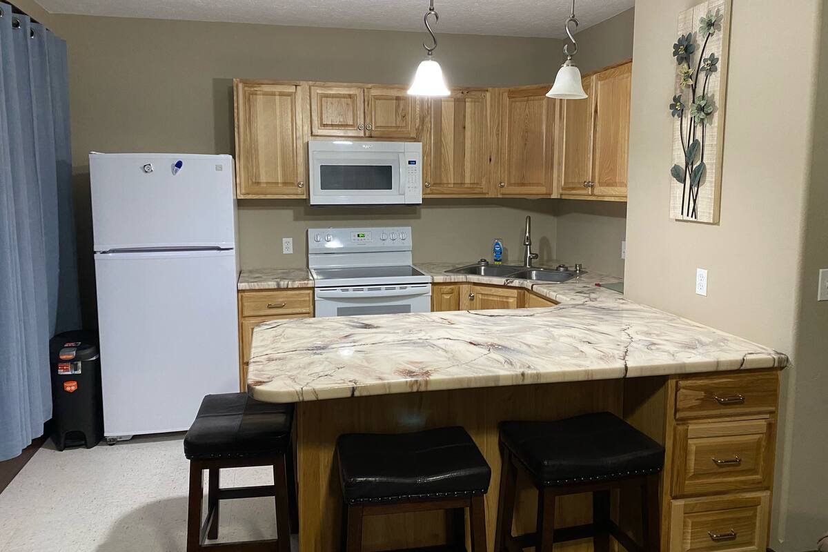 A kitchen with wooden cabinets, a white refrigerator, a microwave, an oven, a sink, and a marble countertop. There are three stools and a small cabinet with drawers.