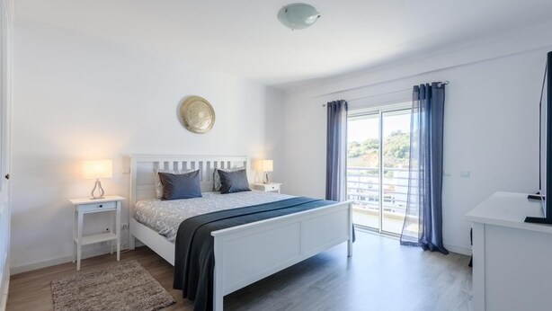 A bedroom with a bed, bedside tables, a lamp, a rug, a ceiling light, and a view through a window with curtains. - Amazing Apartment in Albufeira (Albufeira)