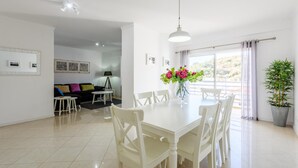 A dining area with a white table and chairs, a vase with flowers, and a view of the outdoors through large windows. - Amazing Apartment in Albufeira (Albufeira)