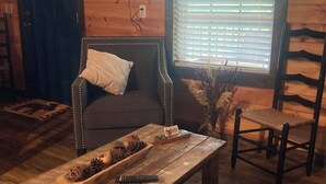 L’intérieur d’un chalet chaleureux, avec un plancher en bois, un canapé, une table basse en bois ornée de pommes de pin, un panneau et une fenêtre avec des stores.