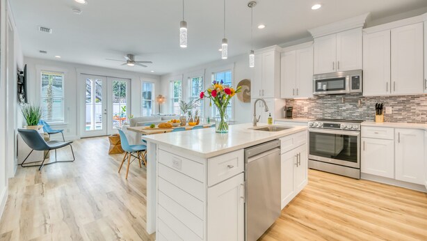 A modern kitchen with white cabinets, a central island, stainless steel appliances, and a dining area with blue chairs. - Joyful Sound by AvantStay | Walk to the Beach (Panama City Beach)