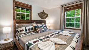 A neatly made bed with a forest-themed bedspread, two bedside lamps, and a wooden headboard. - EV Cottage, Fenced Yard, Pets Free (Sevierville)