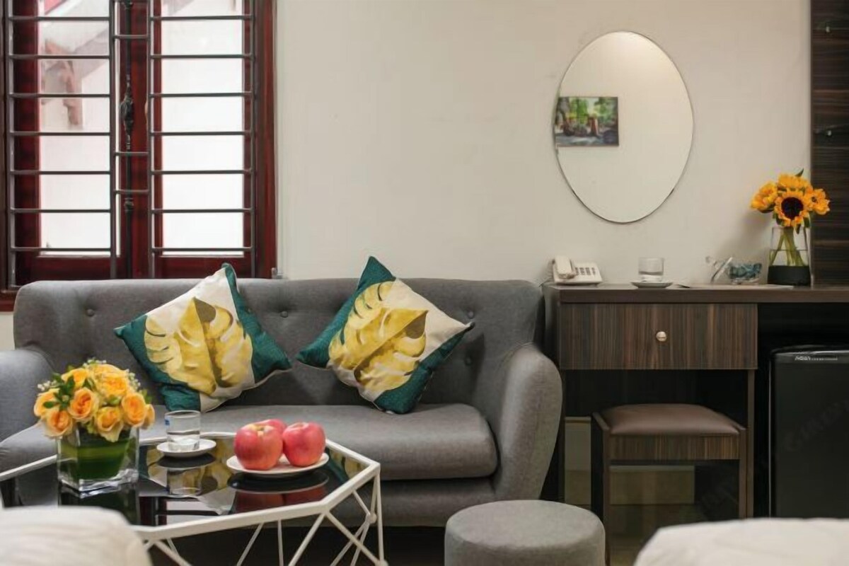 Ein modernes Hotelzimmer mit grauem Sofa, einem kleinen Tisch mit Blumen, Äpfeln und einem Glas Wasser. Ein Schreibtisch mit Telefon und einem Strauß Sonnenblumen ist ebenfalls vorhanden.