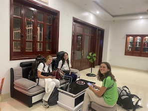 A room with a sofa, a table, and a fan. There are three people sitting on the sofa and one person sitting on the floor. - Hanoi Airport Suites Hostel & Travel (Hanoi)
