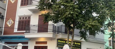 Un edificio de varios pisos con un letrero que dice "Hanoi Airport Suites" y un letrero que indica "Hostel". Un coche rojo está estacionado adelante.