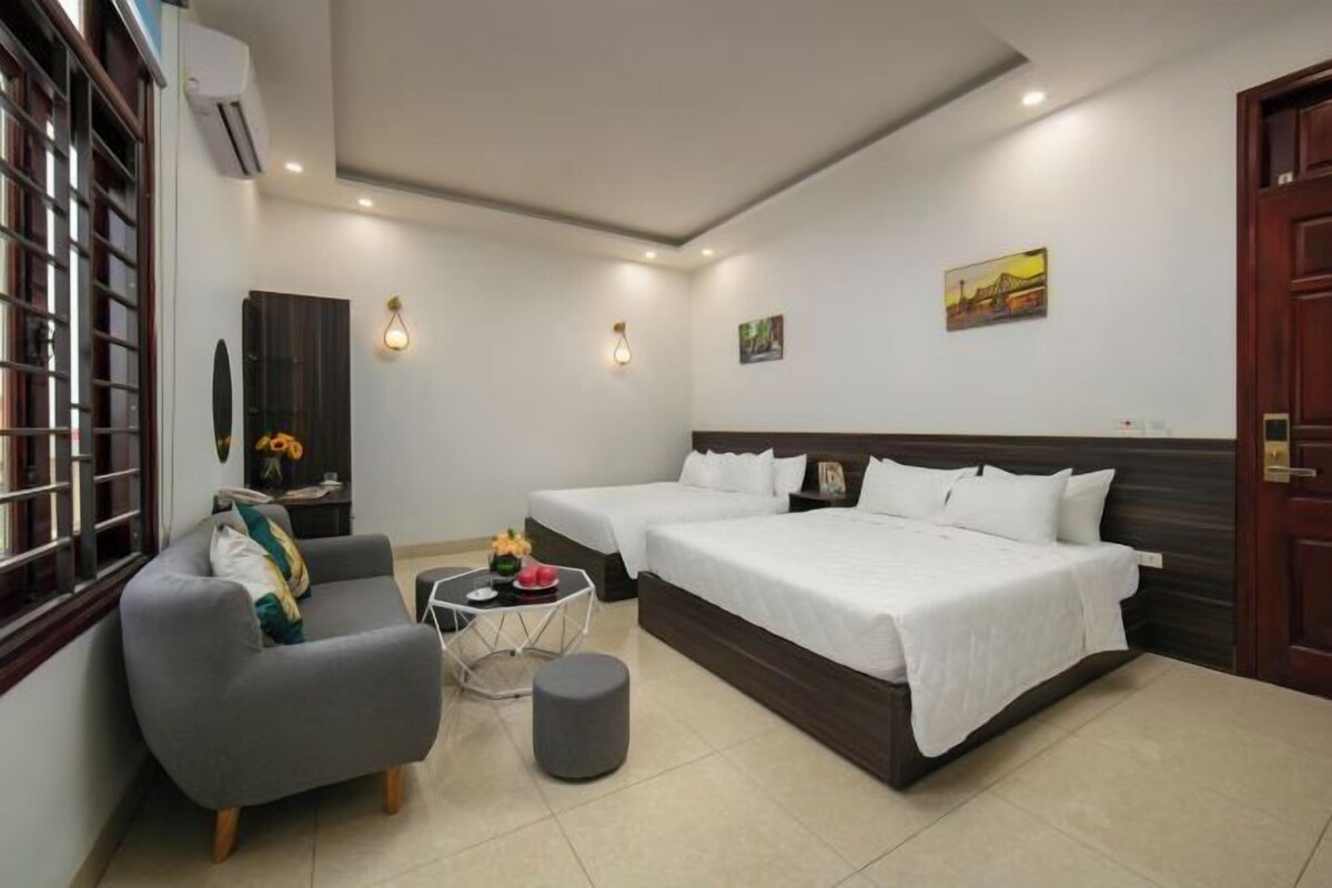 A hotel room with two beds, a sofa, a small table with a vase, and a window with blinds.