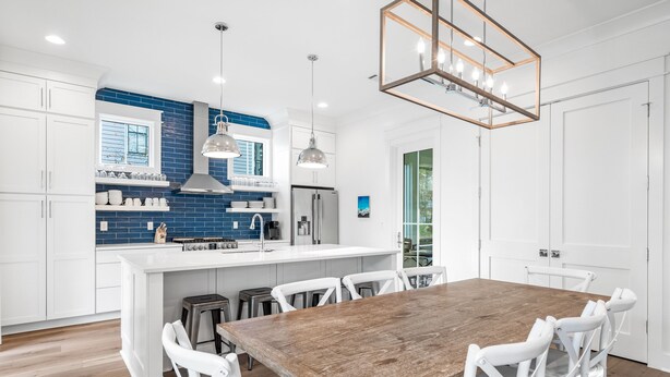 A modern kitchen with white cabinetry, blue backsplash, and a wooden dining table with white chairs.