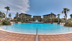 A large outdoor swimming pool with a fountain, surrounded by palm trees and modern buildings.
