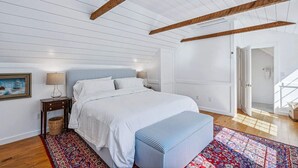 A bedroom with a bed, bedside tables, a nightstand, a patterned rug, and a door leading to another room. - Hillside Farmhouse, Updated & Pet-Friendly (Hartford)