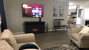 A living room with a fireplace, a flat-screen TV mounted on the wall, and a dining area in the background. - Cozy 2BR Lincoln House (Enid)