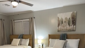 A bedroom with two beds, a ceiling fan, a painting, and a small table with a lamp. - Cozy 2BR Lincoln House (Enid)