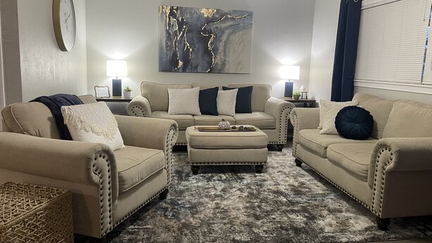 A living room with a beige sofa set, a coffee table, and a rug. There is a painting on the wall and a clock on the wall. - Cozy 2BR Lincoln House (Enid)