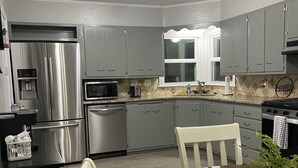 A modern kitchen with grey cabinets, stainless steel appliances, a dining table with chairs, and a potted plant. - Cozy 2BR Lincoln House (Enid)
