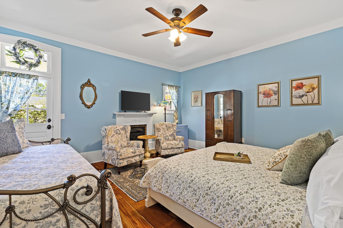 A bedroom with two beds, a ceiling fan, a fireplace, a television, and floral artwork on the walls.