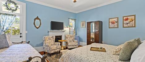 A bedroom with two beds, a ceiling fan, a fireplace, a television, and floral artwork on the walls.