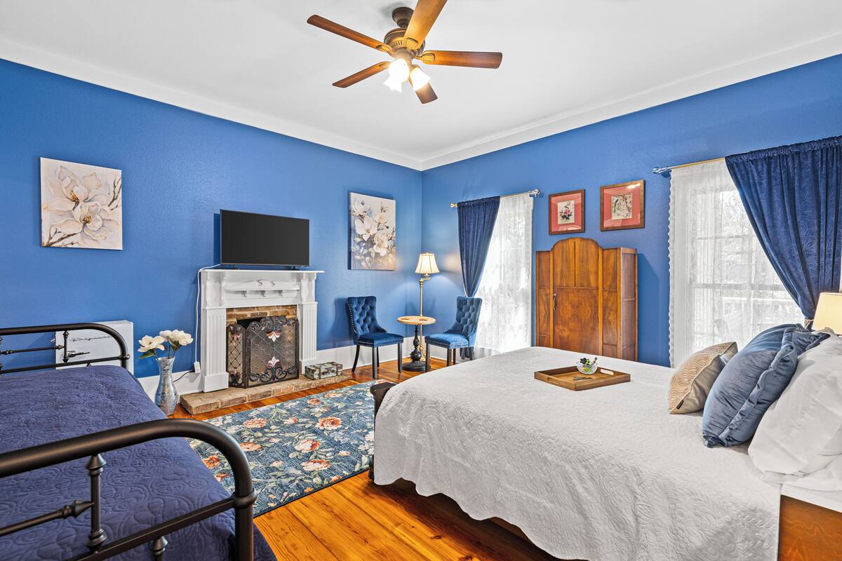 A bedroom with a blue wall, a ceiling fan, a fireplace, a bed, a TV, and a window with curtains.