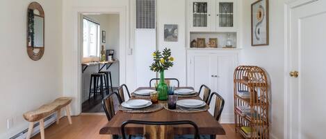A dining room with a wooden table set for four, surrounded by chairs. There is a bench to the left, a bookshelf to the right, and a door leading to another room.
