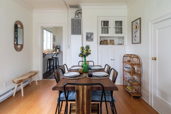 Uma sala de jantar com uma mesa de madeira posta para quatro pessoas, cercada por cadeiras. Há um banco à esquerda, uma estante à direita e uma porta que dá para outro cômodo.