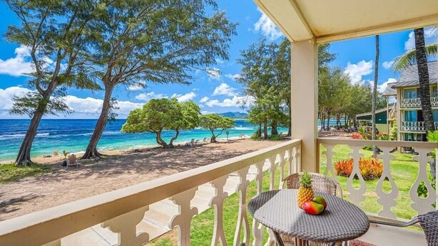 A balcony with a view of the ocean and trees, a table with fruits, and a building in the background. - Islander on the Beach 254 Studio Oceanfront Condo With AC (Kapaa)
