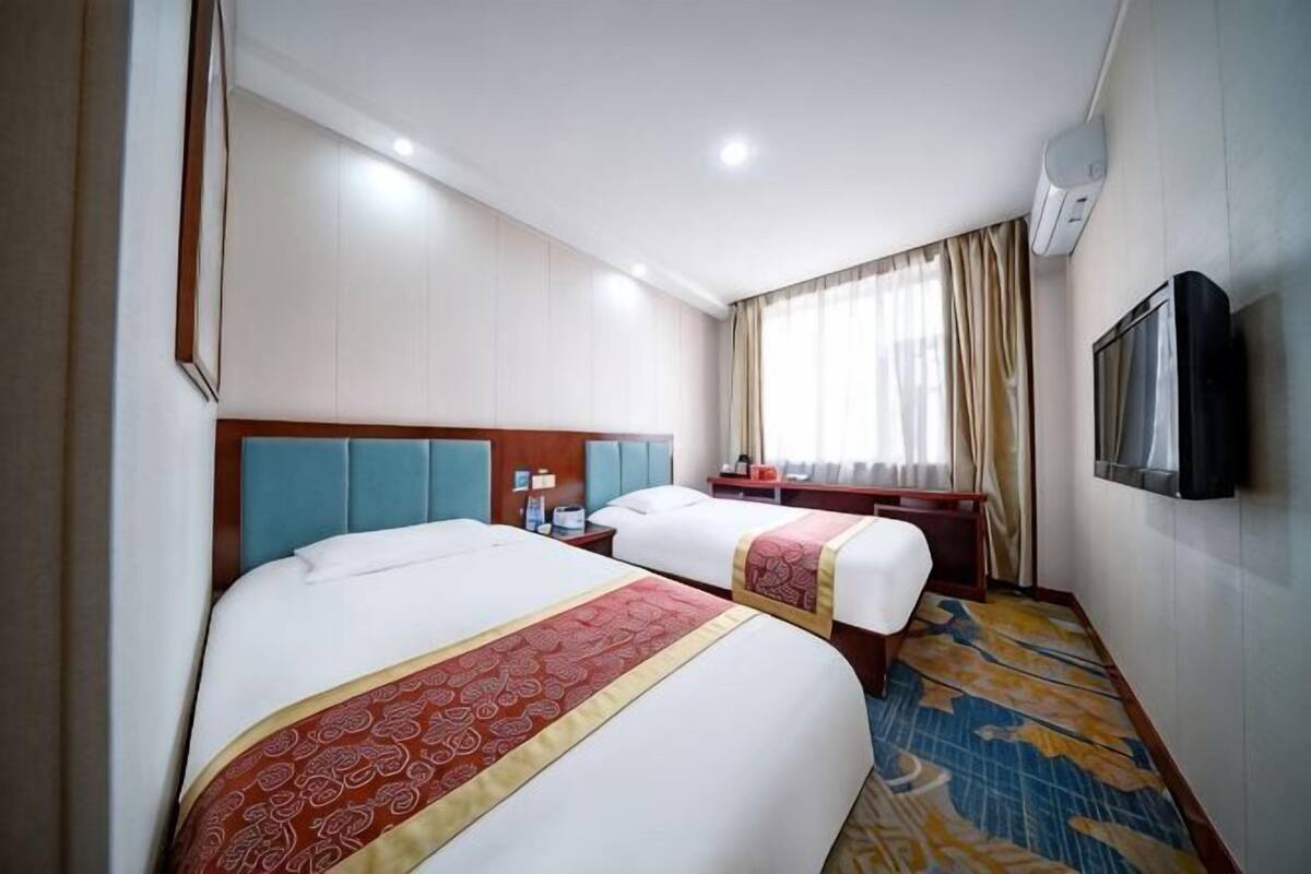 Ein Hotelzimmer mit zwei Betten, einem Fernseher, einem Schreibtisch und einem fensterbelaubten Fenster.