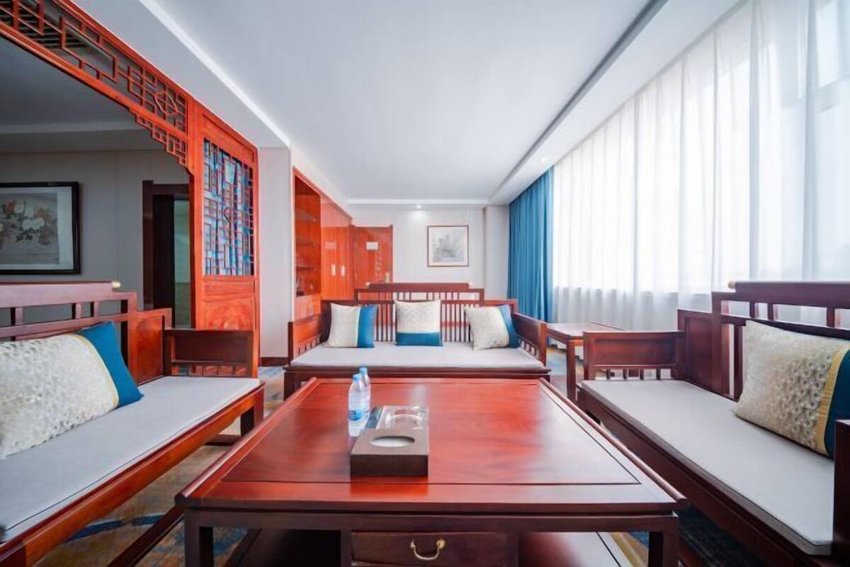 Ein Hotelzimmer mit einer Holz-Sofagarnitur, einem Couchtisch in der Mitte und einem großen Fenster mit Vorhängen.