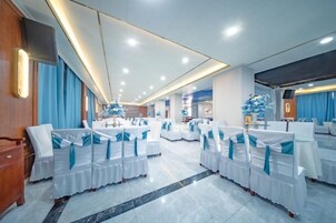 A spacious banquet hall with round tables and chairs, a stage, and a modern ceiling design.