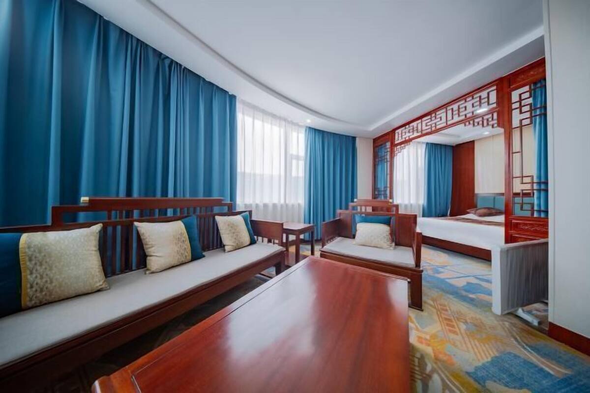 Ein Hotelzimmer mit einem hölzernen Bett, einer Couch, einem Esstisch und einem großen Fenster mit blauen Vorhängen.
