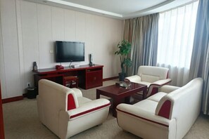 Une chambre d’hôtel moderne dotée d’un téléviseur à écran plat, d’une table basse en bois, de deux canapés blancs agrémentés de touches de rouge, d’une plante en pot et d’une grande fenêtre avec des rideaux.