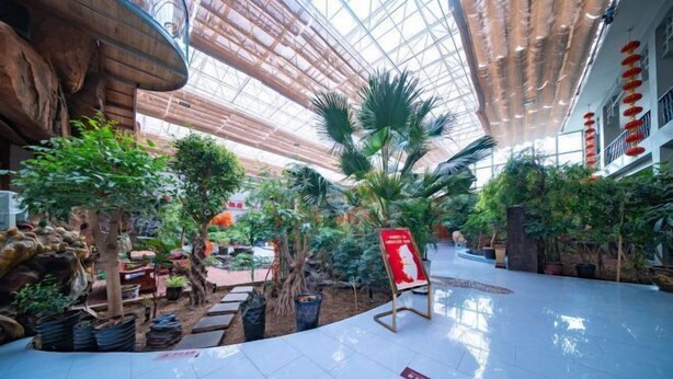 Un vaste atrium intérieur doté d’un haut plafond, agrémenté de diverses plantes en pot et d’un grand panneau rouge placé au centre.