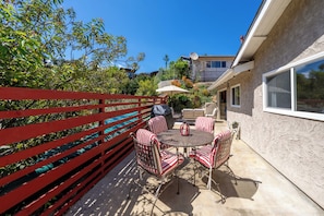Eine Terrasse mit rundem Tisch und Stühlen, ein roter Zaun und ein Gebäude im Hintergrund.