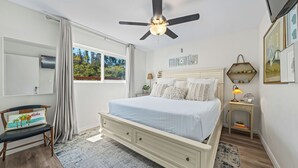 Une chambre à coucher comprenant un lit, un ventilateur de plafond, une table de chevet, une lampe et une fenêtre avec des rideaux.