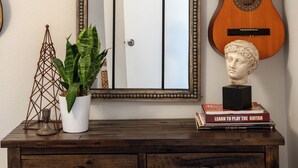 Un miroir avec un cadre décoratif, une guitare, une sculpture en buste, un chapeau et une plante, le tout posé sur une commode en bois.
