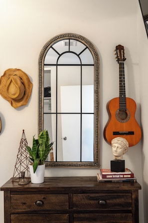 Un espejo con marco decorativo, una guitarra, un busto, un sombrero y una planta sobre un aparador de madera.