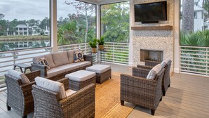 A spacious outdoor deck with wicker furniture, a stone fireplace, and a large TV mounted on the wall. - Lake Views  + Porch Movies, Game Nights and Hot Tub (Bluffton)