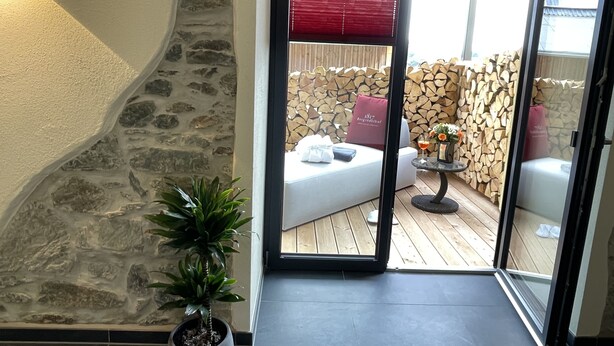 Una habitación acogedora con pared de piedra, una planta en maceta y un ventanal corredero que da a un área de estar exterior con terraza de madera y un toldo rojo.