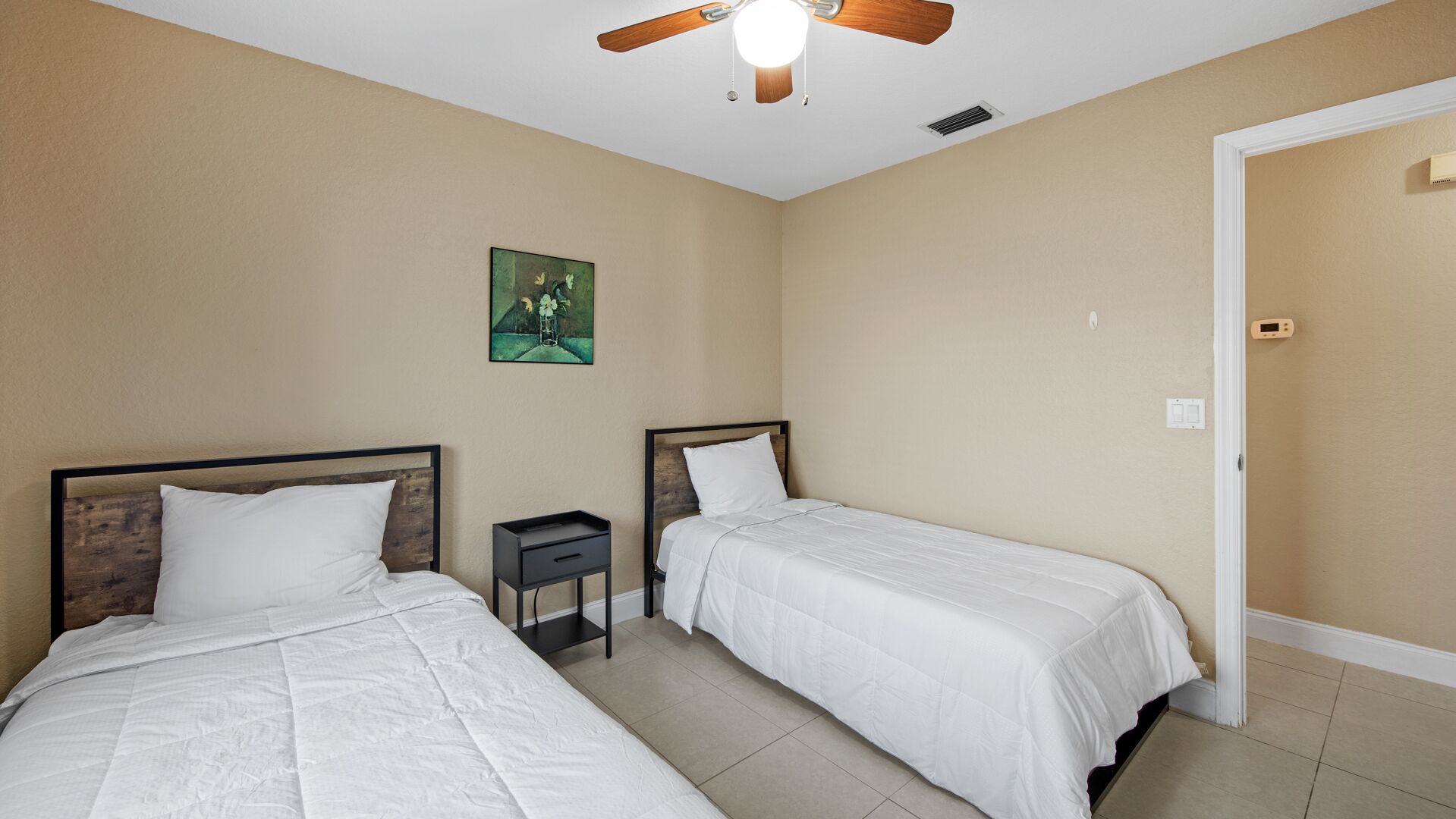A hotel room with two beds, a ceiling fan, a small bedside table, and a door.
