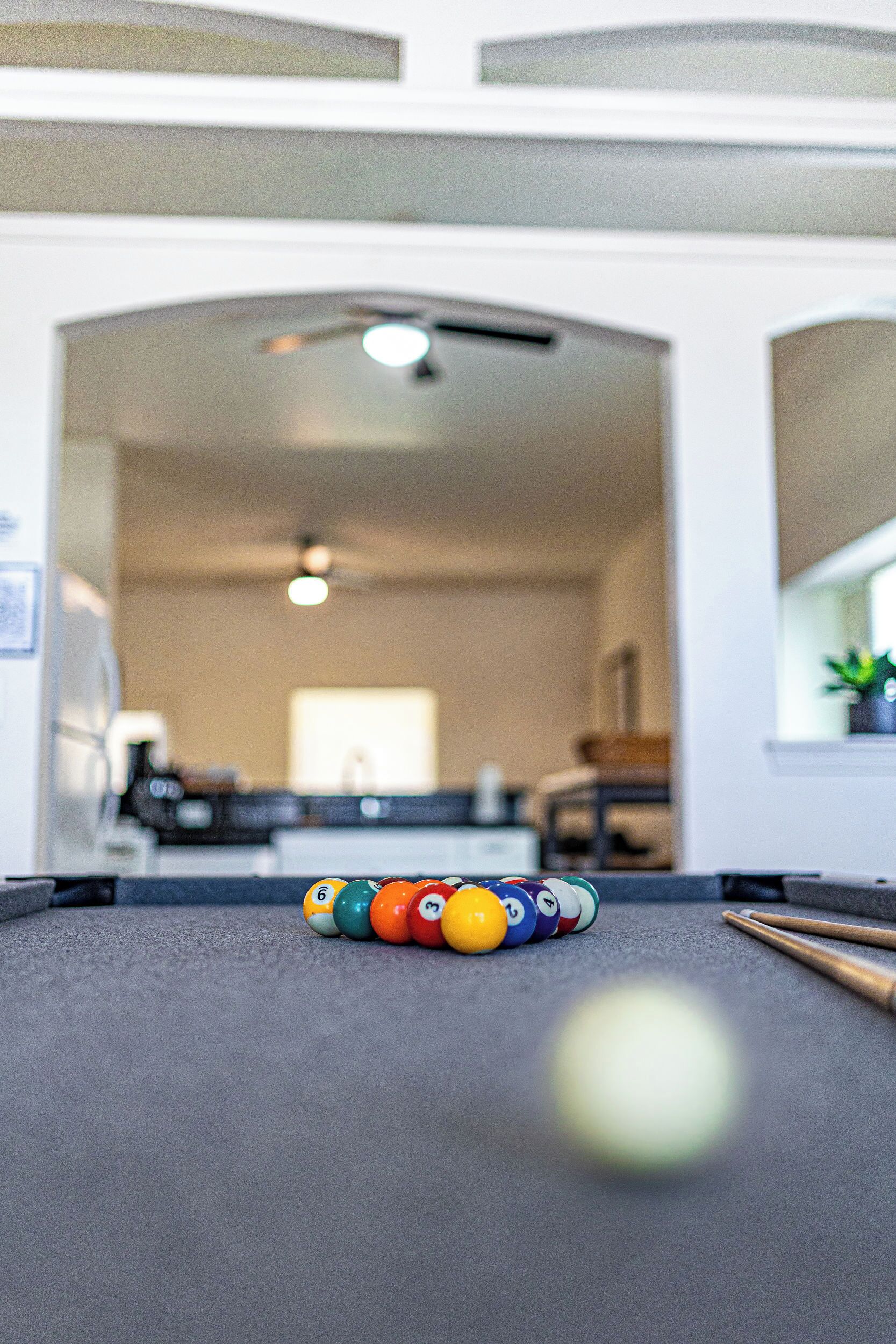A pool table with billiard balls and cues in a room with a kitchen in the background.