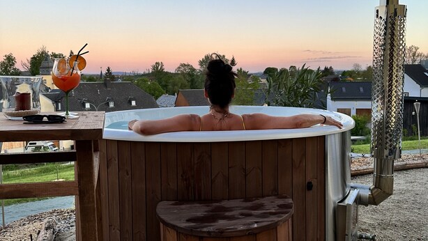 A hot tub with a person relaxing in it, surrounded by a wooden deck and a patio area with a table and drink.