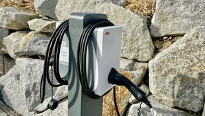An electric vehicle charging station with a label, set against a backdrop of large rocks and a wooden structure in the distance.
