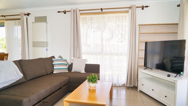 Uma sala de estar com um sofá marrom, uma mesa de centro de madeira, uma televisão sobre um suporte branco e uma janela com cortinas.