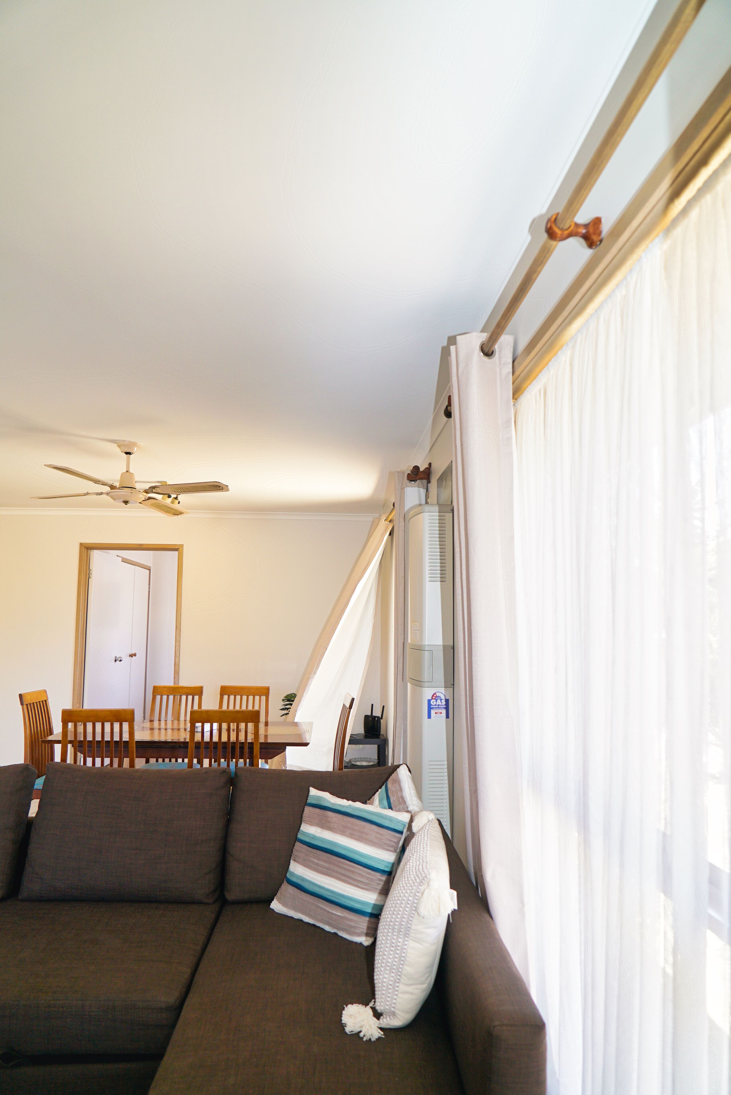 A living room with a brown sofa, a dining table with chairs, a ceiling fan, and a window with curtains.