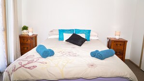 Cama bem arrumada com colcha florida, duas toalhas azuis e uma almofada preta. Há duas mesas de cabeceira com luminárias e uma pequena planta em uma delas.