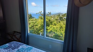 A bedroom with a large window offering a view of a green landscape and the sea.
