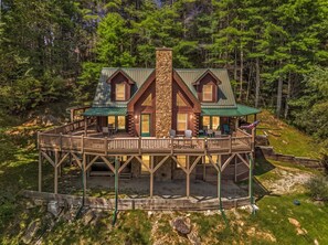 Eine Holzhütte mit einem zentralen Steinkamin, einer zweistöckigen Terrasse und einer Veranda mit Gartenmöbeln.