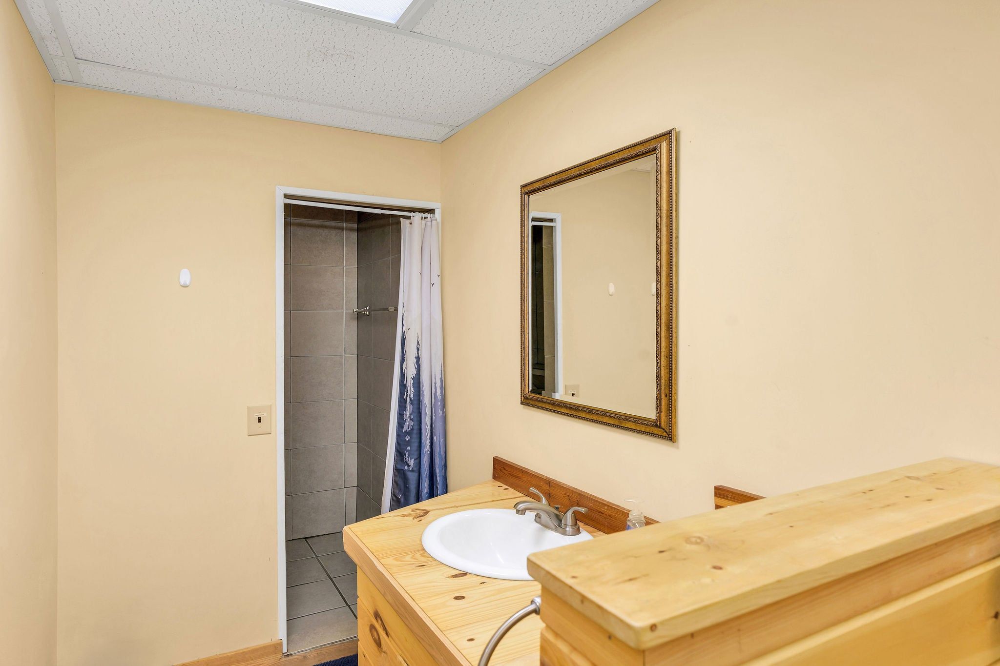 Un baño con tocador de madera, un lavamanos blanco, un espejo con marco decorativo y una ducha con cortina.