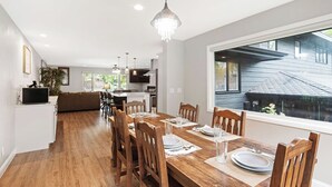 A modern dining area with a wooden table set for four, surrounded by wooden chairs. A chandelier hangs above the table, and a large window provides a view of the outside. - Pets & Kids Love this Montana Dream Home! (Billings)